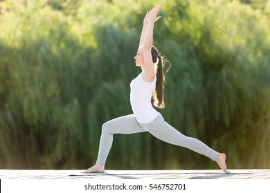 Sporty attractive young woman practicing yoga, standing in Warrior one exercise, Virabhadrasana 1 pose, working out, wearing sportswear, outdoor full length, street background