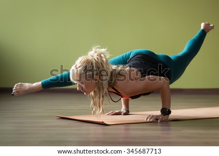 Sporty beautiful blond woman in sportswear working out indoors, doing handstand, standing on orange eco mat in variation of Eka Pada Koundinyasana 2, full length