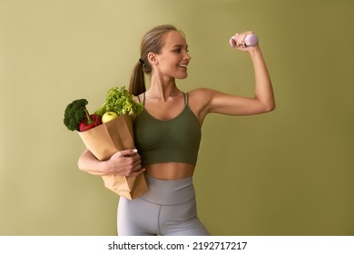 Sporty beautiful woman holding a bag of vegetables and a dumbbell on a green background. The concept of sports and healthy eating. - Shutterstock ID 2192717217