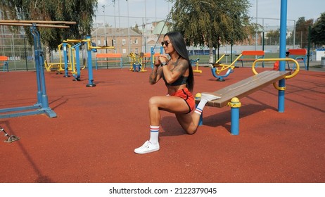 Sporty beautiful woman with long black hair doing bulgarian split squat on the street. Training on the sports ground in the summer in a sports top and shorts