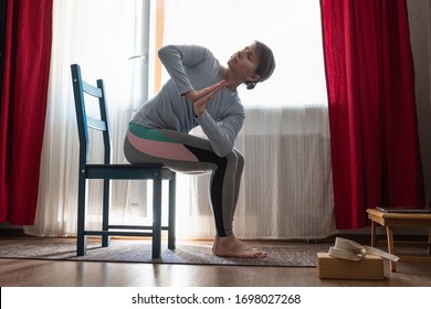 Sporty beautiful young woman practicing yoga using chair, doing Revolved Chair Pose, Parivrtta Utkatasana, working out at the living room at home