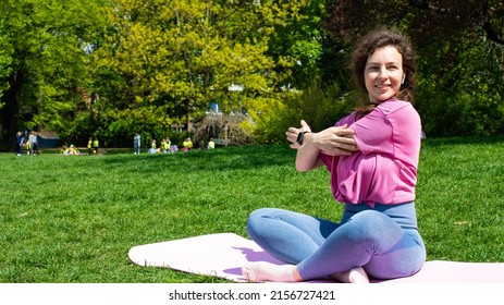 Sporty beautiful young woman stretching arms during exercising outdoors before workout, sitting on mat in city park on green grass in sunny day. Preparing muscles and ligaments. Copy space for text