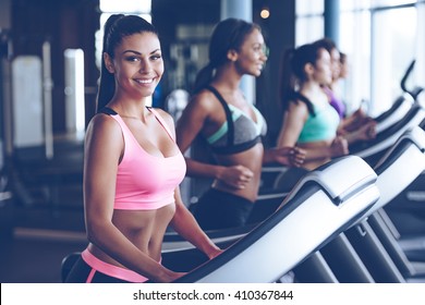 Sporty Beauty. Beautiful Young Cheerful Woman In Sportswear Looking At Camera With Smile While Running On Treadmill At Gym With Other Women