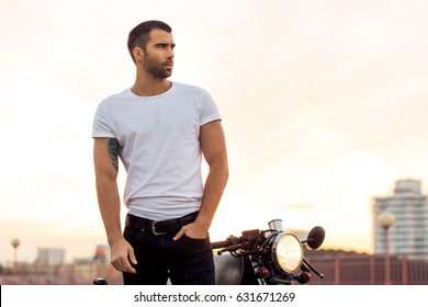 Sporty biker handsome rider male in white blank t-shirt walk away from classic style cafe racer motorbike at sunset. Vintage bike custom made in garage. Brutal urban lifestyle. Outdoor portrait.