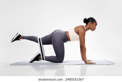 Sporty black lady in fitwear doing quadruped hip extensions in all four position, having workout with rubber loop on fitness mat in white studio. Side view shot. Healthy training routine