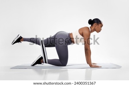 Sporty black lady in fitwear doing quadruped hip extensions in all four position, having workout with rubber loop on fitness mat in white studio. Side view shot. Healthy training routine