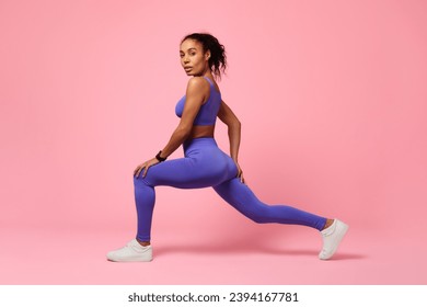 Sporty black woman lunging forward exercising during workout over pink studio background, looking at camera with confidence. Gym training for muscles flexibility. Side view shot - Powered by Shutterstock