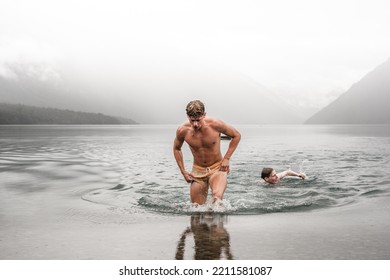 sporty caucasian boy without clothes and with a swimsuit getting out of the water wet and tired with his friend swimming in the cold waters of the lake on a cloudy day with a lot of fog, nelson lakes - Shutterstock ID 2211581087