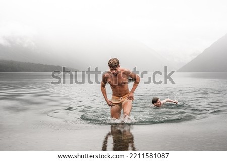 sporty caucasian boy without clothes and with a swimsuit getting out of the water wet and tired with his friend swimming in the cold waters of the lake on a cloudy day with a lot of fog, nelson lakes