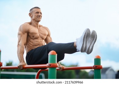 Sporty Caucasian Man With Bare Torso Lifting Legs Up While Training On Parallel Bars Outdoors. Concept Of People, Bodybuilding And Leisure Activity.