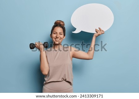 Sporty concept. Studio photo of young happy smiling slim European female wearing beige tracksuit standing isolated in centre on blue background holding black dumbbell and speech bubble with space