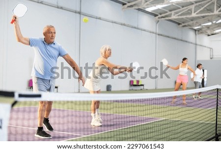 Sporty couple of senior woman and man playing doubles pickleball on indoor court, ready to hit ball. Sport and active lifestyle concept..