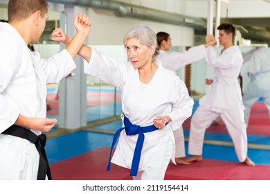 Sporty determined elderly woman in kimono practicing martial arts techniques paired with experienced trainer in training room