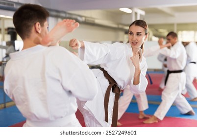 Sporty determined young woman in kimono practicing kicking technique paired with male partner in martial arts training room - Powered by Shutterstock