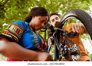 Sporty and eager black woman in yard repairing bike derailleur for summer recreational cycling. Healthy committed african american female cyclist assisted by caucasian man in repairing of bicycle - Powered by Shutterstock