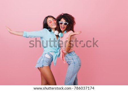 Sporty european woman with bronze skin dancing with cheerful african friend. Indoor photo of happy two girls in summer attires fooling around together.