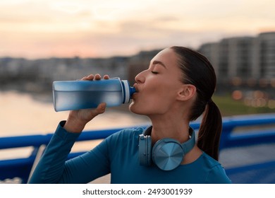 Sporty European woman resting after run and drinking water, standing on city bridge during evening workout - Powered by Shutterstock