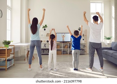 Sporty Family Doing Physical Exercises At Home During A Fitness Workout With An Online Gym Trainer. Mum, Dad, And Kids Standing In The Living Room And Watching A Video Lesson On TV Screen, Back View