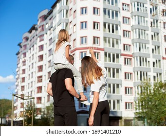 Sporty family of three are standing with their back to the camera and looking at a big block of flats, girl is sitting on father's shoulders. Mother is pointing on a building with big block of flats