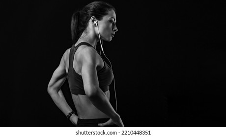 Sporty Female Athlete Listening Music In Headphones In Sportsbra Standing In Power Pose Holding Hands On Waist On Black Background With Empty Copy Space. Back View. Black And White