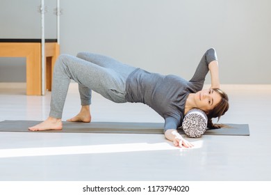 Sporty Female In Gray Sportswear Performing Self-massage Workout With Foam Roller, Lying On Yoga Mat In Pilates Studio. Treatment Of Back An Neck Pain Concept.