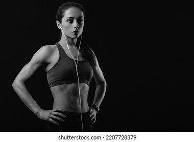 Sporty Female Model Listening Music In Headphones In Sportsbra Standing In Power Pose Holding Hands On Waist Isolated On Black Background With Empty Space. Healthy Lifestyle Portrait. Black And White
