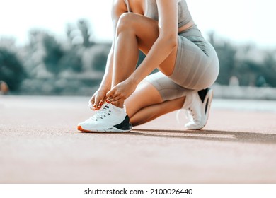 Sporty female tying shoelaces in training. A woman in a sports outfit squats and ties a shoelace. Gray wardrobe and white sneakers at the outdoor training stadium, close shot of a female body - Powered by Shutterstock