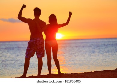 Sporty fitness couple cheering at beach sunset. Happy romantic fit young couple enjoying sunset with arms raised up flexing muscles together. People on sports vacation getaway.