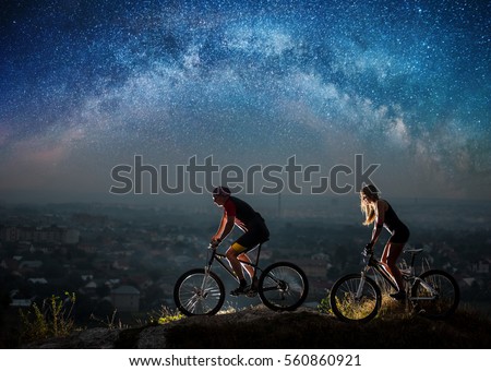 Sporty friends riding a bicycles on the mountain hill at night under starry sky and Milky way. Below is a city in the distance