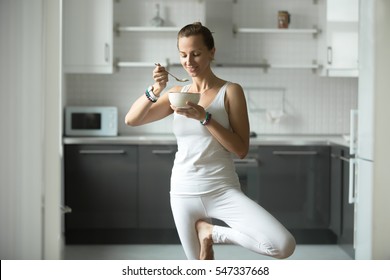 Sporty funny attractive woman having lunch in kitchen while practicing yoga, standing in Tree exercise, Vrksasana pose, work out in white sportswear, enjoying food, indoor, home interior background