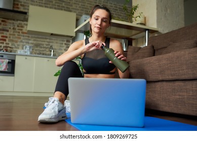 A Sporty Girl In A Black Workout Tight Suit Is Opening A Bottle During Watching An Online Video On A Laptop. A Coach Conducting A Virtual Fitness Class On The Blue Yoga Mat At Home.