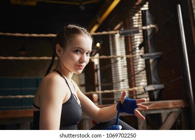 Sporty girl with braids sitting indoors, wrapping bandages on her hands, preparing for fight. Beautiful young sportswoman in black top smiling at camera, tying hand wraps before boxing training in gym - Powered by Shutterstock