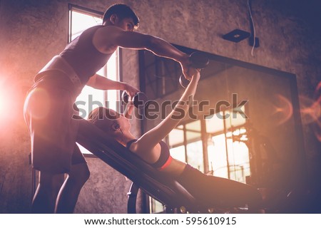 Sporty girl doing weight exercises with assistance of her personal trainer at public gym.