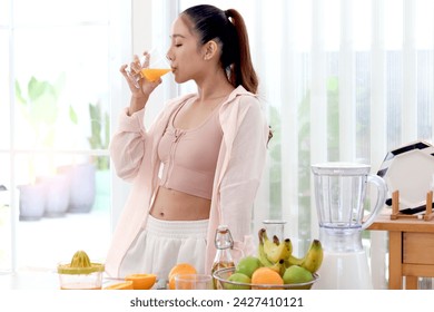 Sporty girl drinking orange juice while standing at kitchen. Beautiful happy Asian woman with fruit basket, healthy smoothie ingredients and mixing blender on wooden table at home kitchen. - Powered by Shutterstock