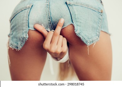 Sporty girl in jeans shorts with fit ass show middle finger, fuck you off sign between her legs in front of a white wall. Outdoor funny lifestyle portrait, concept of brutal, rebel, punk, indecent