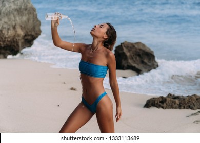 Sporty girl in swimsuit at beach pours clear water on herself from bottle. Beautiful woman drinks cool water on hot day. Concept bikini, summer, travel, swimwear, body, vacation, cool drinks