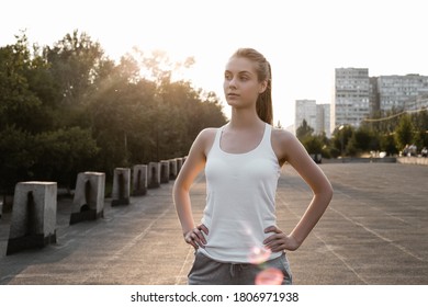 Sporty girl in white tank top stands with her back to the camera and with hands on hips.