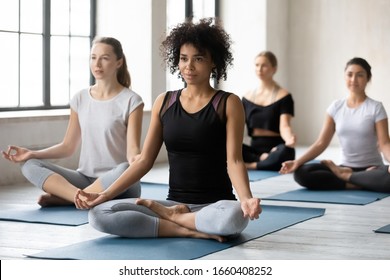 Sporty girls and guys sitting in lotus pose during yoga class led by african woman coach. Group of people do meditation practice, improve inner harmony, healthy lifestyle, mind body relaxation concept