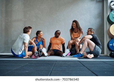 Sporty Group Of Young Friends Talking And Laughing While Sitting Together On The Floor Of A Gym After A Workout