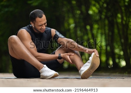 Sporty handsome african american man got injured while training outdoors, sitting on yoga mat at public park, massaging his ankle, suffering from pain, panorama with copy space