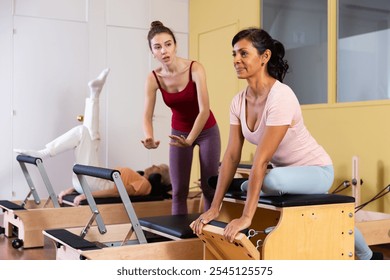 Sporty Hispanic woman practicing Pilates system exercises on wunda chair under guidance of positive qualified young female trainer - Powered by Shutterstock