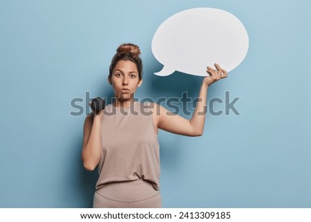 Sporty lifestyle. Indoor photo of young fit pretty European lady wearing beige tracksuit standing isolated on blue background in centre holding dumbbell and speech bubble with space for promotion