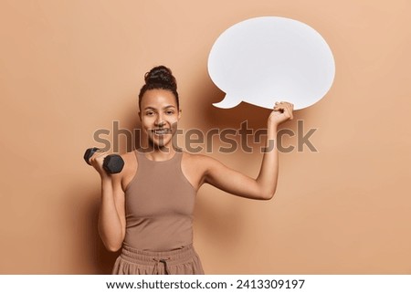 Sporty lifestyle. Indoor shot of young happy pretty smiling fit European girl standing in centre on beige background wearing brown tracksuit doing physical exercise using dumbbell with speech bubble