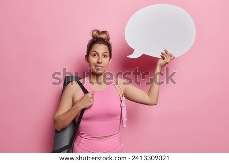 Sporty lifestyle. Studio waist up of European girl with mat and jumping rope wearing tracksuit standing in centre isolated on pink background holding white speech bubble for your advertisement