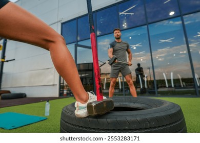 Sporty man in athletic wear is using a sledgehammer to hit a large tire outdoors at a gym, a training partners leg is visible in the foreground. - Powered by Shutterstock