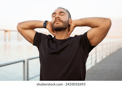 Sporty man in black t-shirt closes his eyes touching neck, having moment of peace, relieving from aching muscles discomfort and neckache after outdoor workout by the sea - Powered by Shutterstock