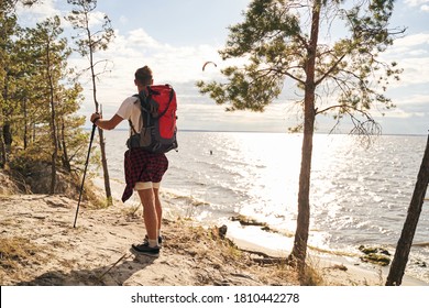 Sporty man is carrying backpack while hiking with trecking sticks in forest near sea shore