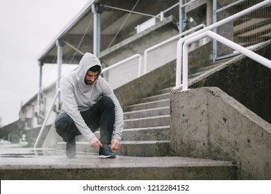 Sporty man getting ready for winter running and outdoor fitness workout under the rain. Sportsman lacing sport shoes on urban stairs.