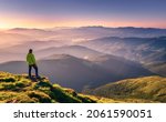 Sporty man on the mountain peak looking on mountain valley with sunbeams at colorful sunset in autumn in Europe. Landscape with traveler, foggy hills, forest in fall, amazing sky and sunlight in fall