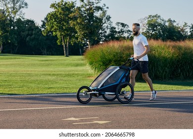 Sporty Man In Sportswear Running With Baby Stroller At Sunny Morning In City Park During Fatherhood Time. Side View